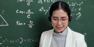 年轻的亚洲女教师手持粉笔，戴着耳机，与学生看着摄像机进行视频会议。女教师在课堂上培训数学黑板在线课程。
