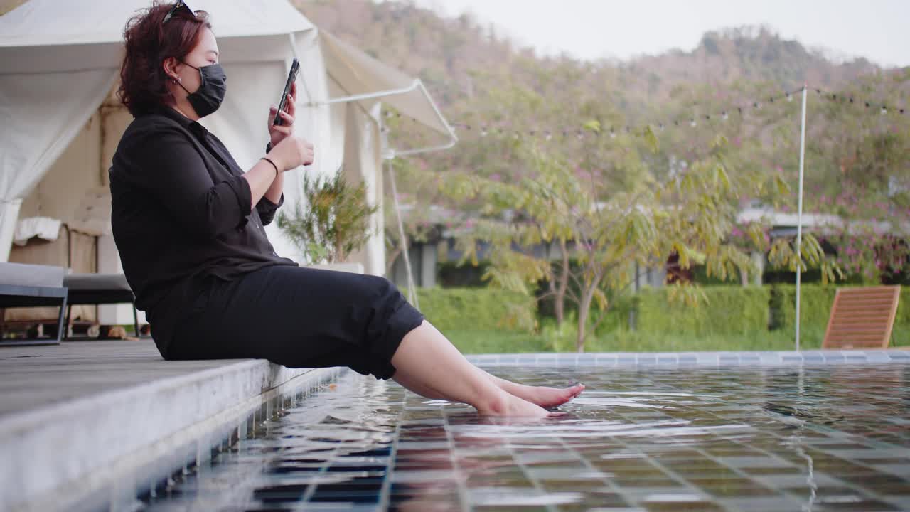 漂亮的年轻亚洲女人保护口罩使用智能手机放松假期假期无忧无虑在泳池与自然背景