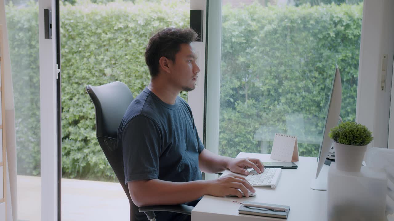 描绘严肃的年轻亚洲自由职业者在家里的电脑上工作。休闲专注的企业家一边坐在家里的办公桌上用电脑工作，一边开发项目