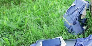 书包，笔记本电脑，电子书，笔记本和牛仔衬衫躺在绿色的草地上。风吹动草