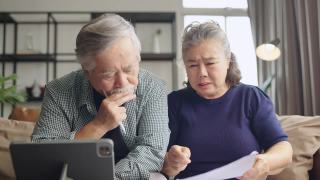 退休的亚洲老夫妇在沙发上一起检查和计算财务账单，参与财务文书工作，在客厅用电子银行笔记本电脑在线纳税视频素材模板下载