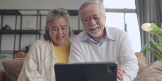 老亚洲老夫妇退休年龄坐在沙发沙发上视频给他们的家人打电话用平板设备社交连接家隔离呆家状态秩序概念，幸福老笑快乐
