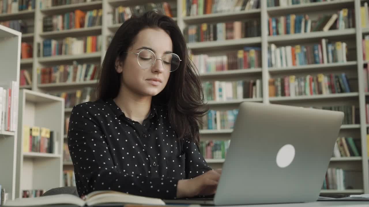 疲惫的女性按摩太阳穴，偏头痛，在电脑工作后头痛，坐在书桌前拿着笔记本电脑在图书馆，疲惫的年轻女性闭上眼睛触摸头部，缓解疼痛