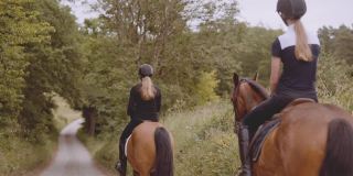 两位女骑手骑着棕色的马在狭窄的道路上