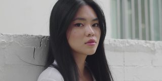 黑色长发的亚洲美女肖像斜倚在白色的墙外。近距离看镜头的酷新潮女孩的脸。迷人的外观