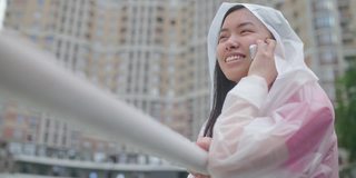 微笑的亚洲女人享受下雨的天气在城市，在室外讲电话