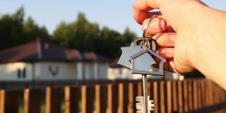 钥匙圈和钥匙链在手里。篱笆和小屋的背景。搬新家，抵押贷款，买房地产，租房预订，梦想住在农村。本空间