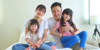 亚洲幸福家庭的肖像微笑和看相机在家里的床上。爱的夫妇和年幼的小女孩兄弟姐妹的女儿喜欢早上在卧室一起活动后从家里醒来。
