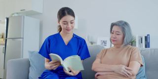 亚洲女护士照顾老年妇女在养老院。年轻的护理治疗师医生坐在沙发上，读一本书给年老的残疾病人快乐。医疗保险服务理念。