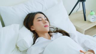 亚洲美丽的女孩穿着睡衣懒洋洋地躺在卧室的床上。迷人的困年轻女性感觉不好发烧和头痛，不想起床后，在家里的清晨。视频素材模板下载