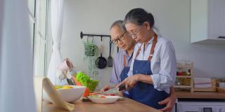 爱亚洲的老夫妇穿着围裙在厨房做饭。英俊强壮的老爷爷奶奶戴着眼镜，在家享受退休生活活动。家庭关系的概念。