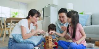 亚洲幸福的一家人坐在地板上，在客厅里一起玩儿童玩具。漂亮的父母和爸爸喜欢和年轻可爱的女儿们在家里玩滚塔木块游戏