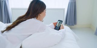 亚洲美丽的女人谈视频电话与男朋友在床上在家里。穿着睡衣的漂亮女孩喜欢早上在卧室醒来后用手机虚拟远程在线与年轻男子聊天