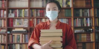积极的亚洲学生戴着口罩，拿着一叠书，在流行病期间教育