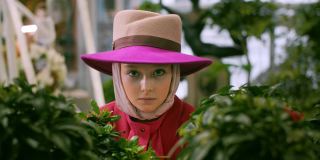 戴着帽子的浪漫女人在橘子园的绿色植物中张望。画一个戴着帽子的漂亮女人在绿色植物丛生的花园里摆姿势。