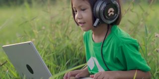 小女孩戴着耳机，在绿草地上用笔记本电脑社交媒体。乡村景色。在夏天的休闲时间