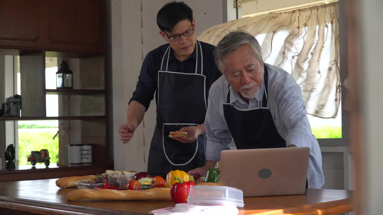 快乐亚的小儿子和老父亲一起在家里的厨房里用笔记本电脑在线烹饪课制作新鲜的蔬菜食品