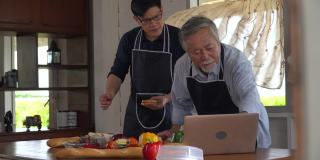 快乐亚的小儿子和老父亲一起在家里的厨房里用笔记本电脑在线烹饪课制作新鲜的蔬菜食品