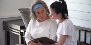 亚洲年轻的女儿和年长的母亲在家里一起读书。年轻女子读人寿保险单给老妈妈听