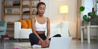 瑜伽教练教练在笔记本电脑上视频通话坐在瑜伽垫上做莲花姿势。健身教练
