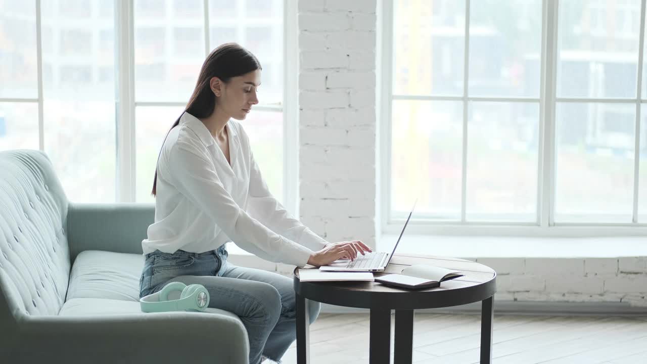 体贴的年轻女商人，黑发，坐在沙发上用笔记本电脑打字。