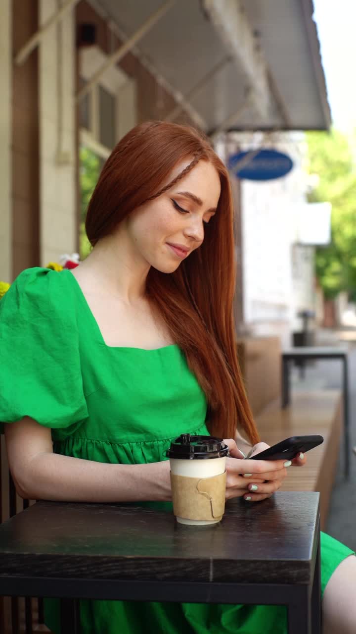 在阳光明媚的夏日，微笑的年轻女子的垂直肖像使用浏览手机坐在餐桌咖啡杯在户外咖啡馆露台。