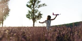女孩拿着飞机在夕阳西下的山坡上奔跑。快乐女孩带着玩具飞机在夕阳下的薰衣草田上奔跑。孩子们玩玩具飞机。概念大孩子梦想