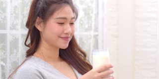 健康亚洲年轻女性在家里喝含钙牛奶强健骨骼，微笑印度女性拿着玻璃豆奶享受营养健康生活，健康与天然牛奶新鲜概念