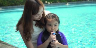 疲惫的女孩在喝着水，幸福的亚洲家庭在酒店的游泳池里玩着，享受着暑假。年轻的母亲抱着女儿一起在户外游泳池里放松