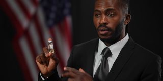一名非洲裔美国男性政客正在为新冠疫苗注射做广告，背景是美国国旗。一个严肃自信的男人的肖像，穿着正式的黑色西装，谈论疫苗接种的需要。