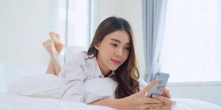亚洲美女早上在家里的床上用手机聊天。快乐随意的漂亮女性躺在卧室享受假日周末，在家里用智能手机和科技交流。