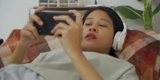 亚洲女人躺着，戴着无线耳机，用智能手机玩游戏