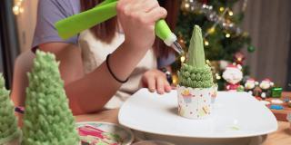 亚洲妇女的手从奶油榨汁机榨取绿色奶油，在自制的圣诞树上做成树叶，作为庆祝圣诞节的甜点，12月假日在公寓里装饰圣诞树，礼盒，圣诞老人。