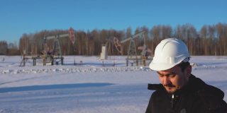 一个戴着白色头盔的工程师在打电话。后面有油泵。俄罗斯冬季石油生产