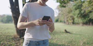 一名亚洲男子身穿白色休闲衬衫，一边走在绿色公园里一边使用智能手机，双手使用无线技术，放松休闲活动，无线上网，在树荫下等待