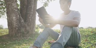 年轻的亚洲人坐在公园大树下，手拿数码笔记本电脑，自由职业者穿着休闲服在户外远程工作，坐在舒适的草地上，镜头光晕背后，自然的人