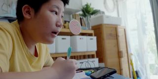 亚洲男孩一边做作业一边在家学习在线课程，社交距离的生活方式概念。