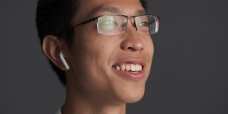 年轻迷人的亚洲男人的肖像戴着眼镜，在灰色的背景下用无线耳机梦幻般的交谈