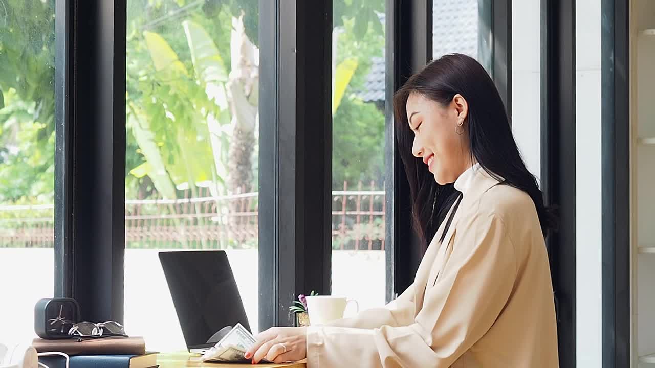 一个亚洲女人坐在咖啡店里，拿着笔记本电脑，喝着咖啡，看着别处。亚洲女性放松在café。