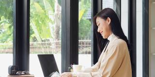 一个亚洲女人坐在咖啡店里，拿着笔记本电脑，喝着咖啡，看着别处。亚洲女性放松在café。