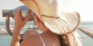 穿着沙滩服戴着沙滩帽的女人用固定的双筒望远镜看。双筒望远镜在海面附近的观察甲板上。在敖德萨港口。码头上的豪华游艇。商用机器，自动售货业务。