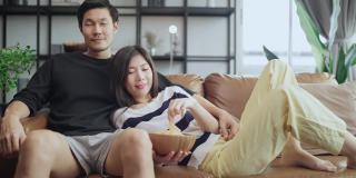 亚洲成人恋人结婚夫妇享受在沙发沙发上看电视，一起在家里享受宁静和幸福，居家隔离的生活方式