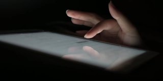 在床上睡觉前，一名女性在不开灯的情况下，手指滑过平板电脑屏幕玩社交媒体。