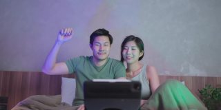 亚洲成人情侣结婚夫妇享受唱歌和幸福舞蹈一起移动手挥手看音乐会在线从笔记本或平板电脑在家里的床上，居家隔离的生活方式