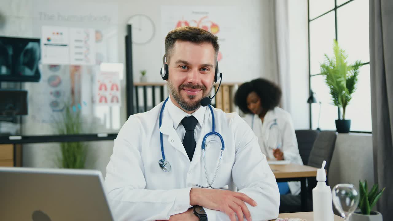 英俊快乐的合格成人胡子医生戴着耳机坐在他的工作地点在医疗办公室和手指对着摄像机