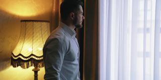旅馆靠窗的男人。穿着白衬衫的年轻商人站在旅馆房间的窗户旁边，透过窗户往外看。后视图。