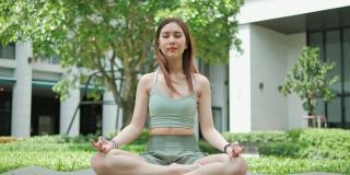 年轻的亚洲女人在运动服坐在瑜伽户外绿色公园。健康，运动，健身和生活方式的概念。瑜伽和冥想对健康有好处