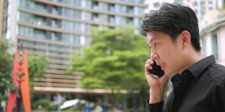 自信的年轻亚洲商人走在户外，在城市街道上用智能手机聊天