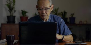 年长的自由职业男性作家戴着眼镜，在晚上用笔记本电脑打字，喝着热咖啡。资深作家在家里的桌子上用电脑工作。居家办公