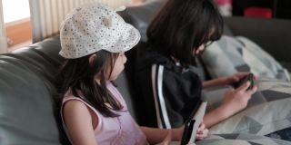 两个亚洲(泰国)女孩穿着便装坐在灰色沙发上，用手机玩着有趣的网络游戏。戴着帽子的妹妹用平板电脑控制屏幕。